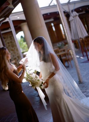Elegant bride in fitted gown and long veil holding white bouquet in outdoor venue.