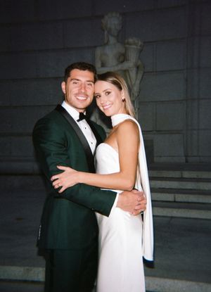 Couple in formal attire embracing in front of classical architecture with a statue.