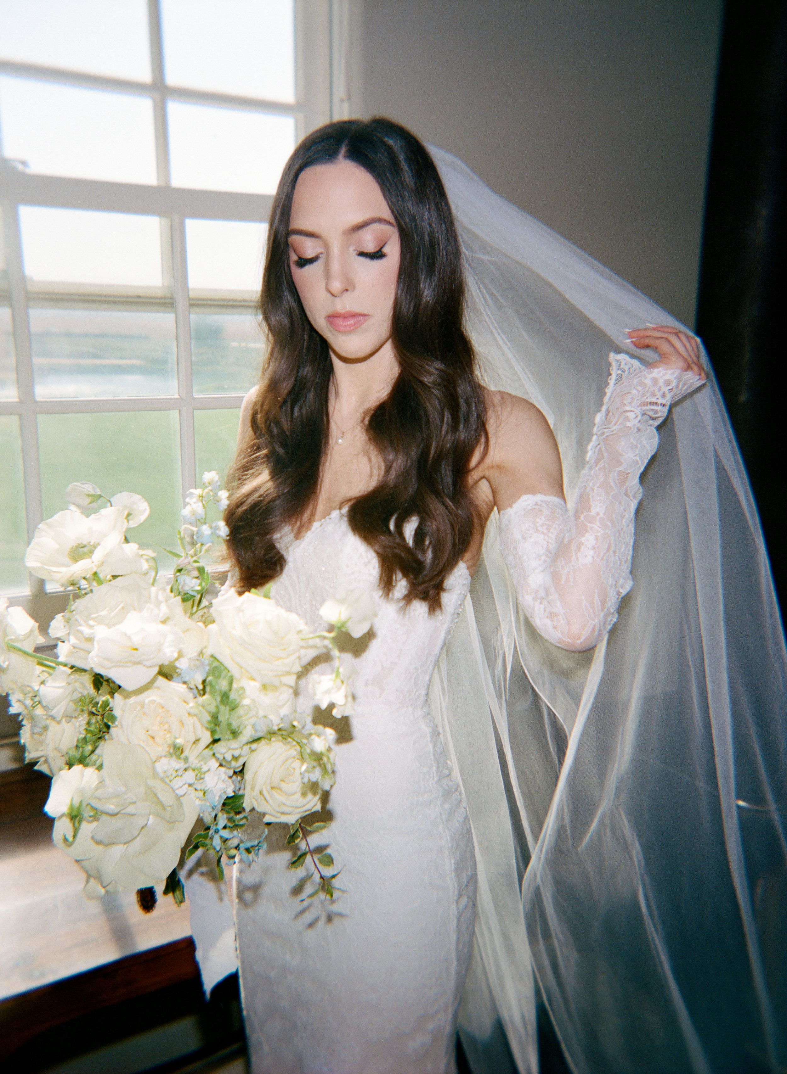 A long white wedding dress and veil captured near a window with white floral bouquet.