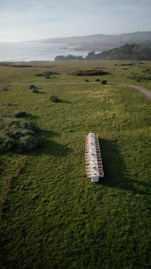 Long dining table set up on a grassy coastal cliff overlooking the rugged California shoreline at sunset.