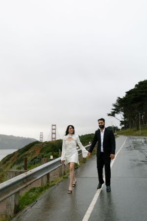 A couple walking along a wet road with the Golden Gate Bridge visible in the background.