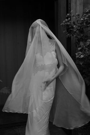 A black and white photograph of a bride in a flowing veil and form-fitting wedding dress standing elegantly.