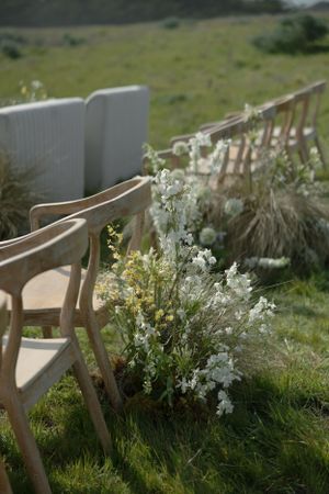 Wooden chairs lined up outdoors with delicate white wildflowers and greenery decorating the aisle for a garden ceremony.