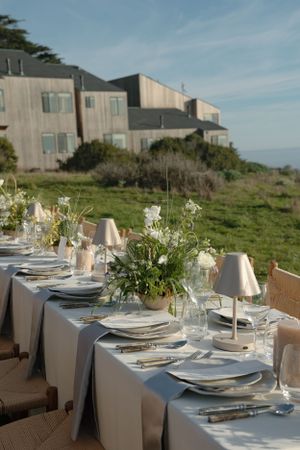 Elegant outdoor dining table set with white linens, floral centerpieces and table settings overlooking coastal architecture.