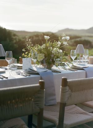 Elegant outdoor table setting with white floral centerpiece and wine glasses against scenic mountain backdrop at sunset.