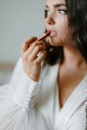 Close-up of woman in white robe applying lip makeup during wedding preparation.