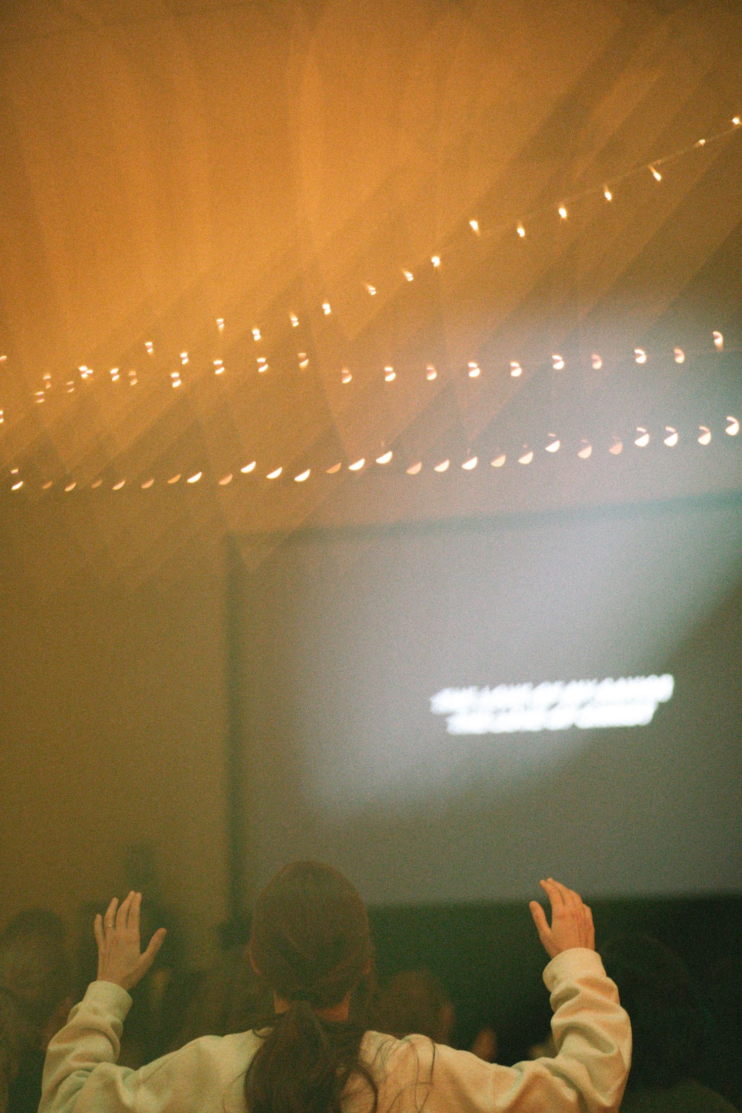 Person raises hands in worship at indoor event with string lights and projection screen.