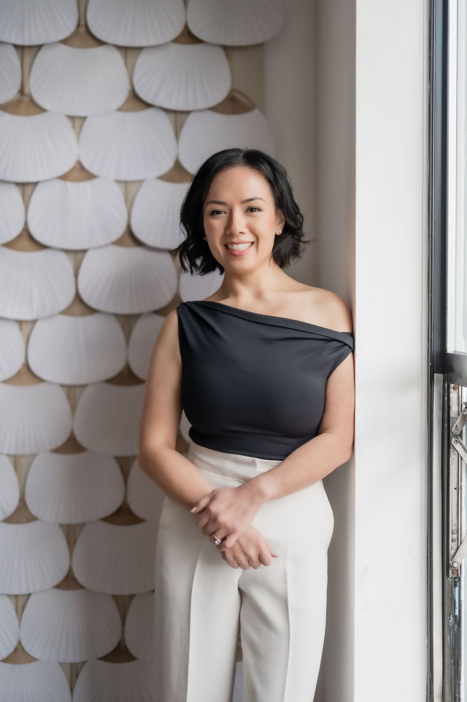Woman in black asymmetrical top and white pants poses against wall with decorative white fan pattern background.