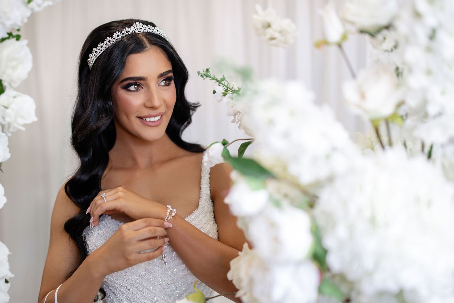Elegant dark-haired bride wearing a crystal tiara and lace gown surrounded by white floral arrangements.