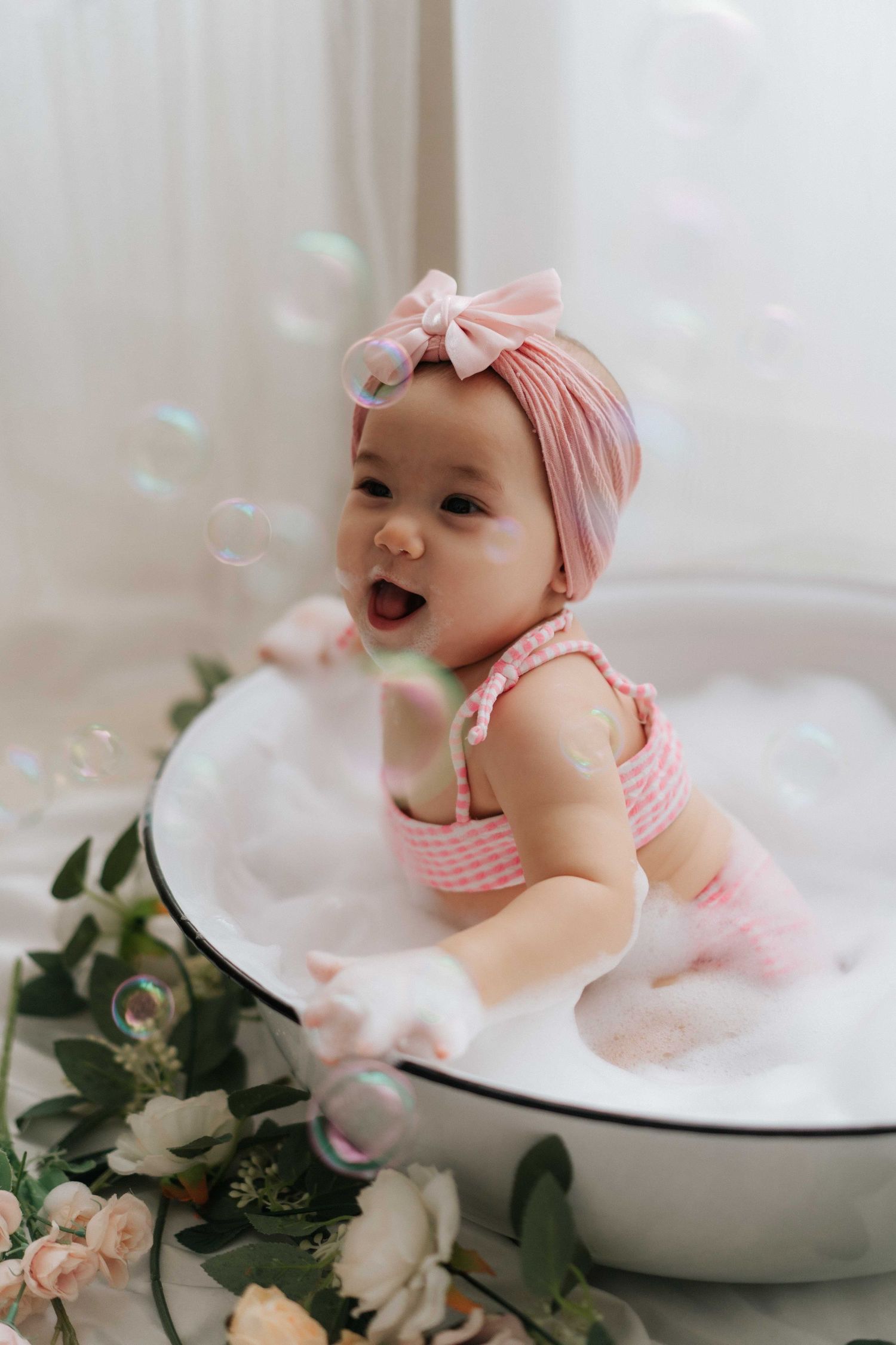 Baby in white overalls sits beside birthday cake with green and white balloon decorations for first birthday celebration.