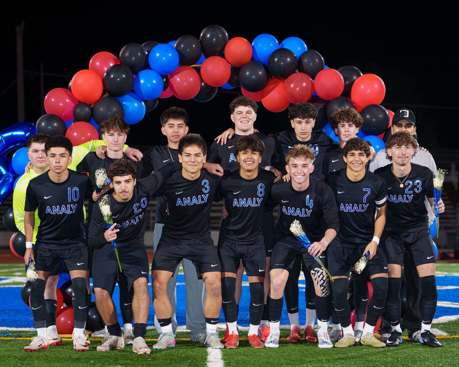 Soccer team poses together in matching navy uniforms under red and blue balloon arch on sports field at night.