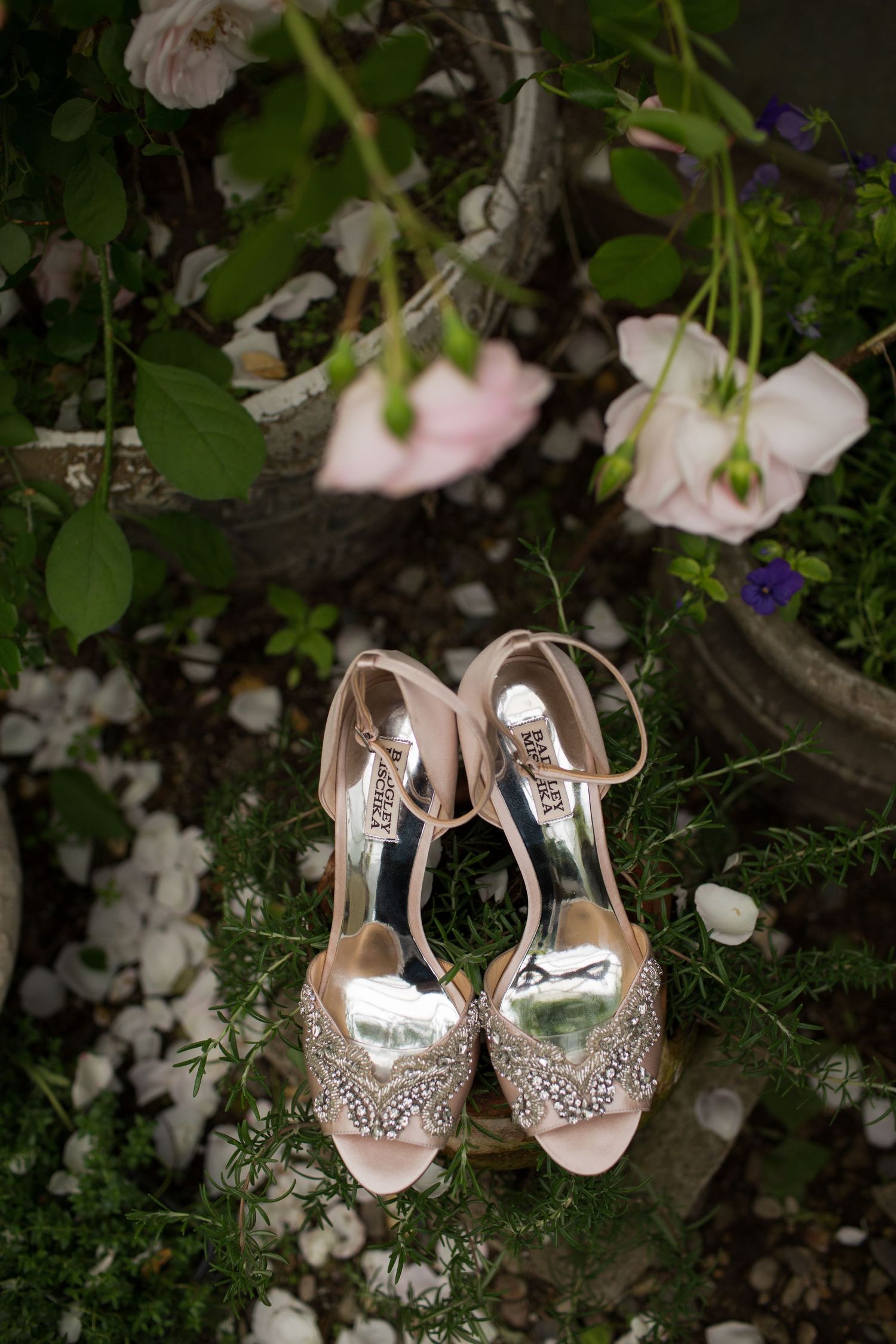 Elegant silver and crystal embellished bridal shoes displayed among white flowers and greenery in a garden setting.