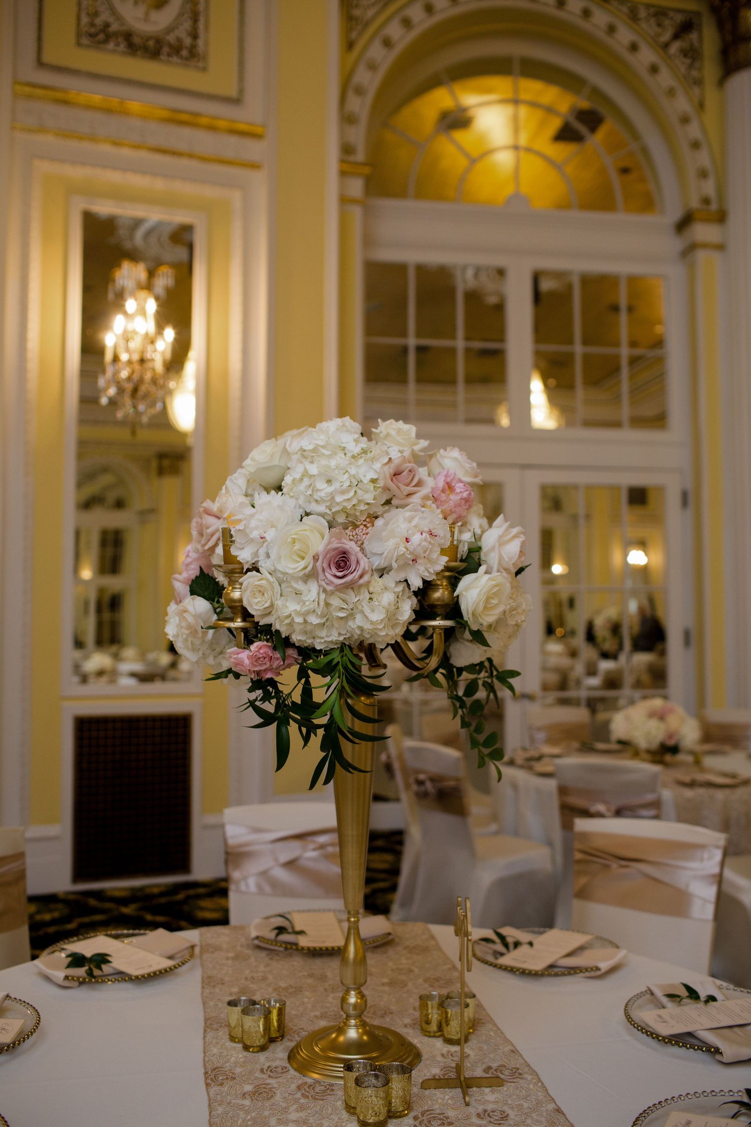 Elegant white and pink floral centerpiece on gold stand in luxurious ballroom with crystal chandelier and arched windows.