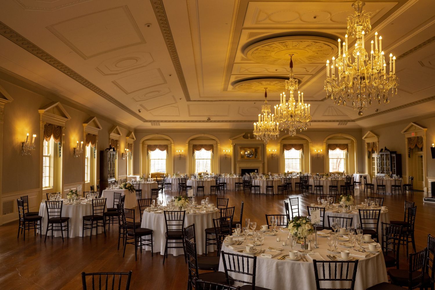 Elegant ballroom with crystal chandeliers, ornate moldings, and round tables set for a formal event in warm lighting.