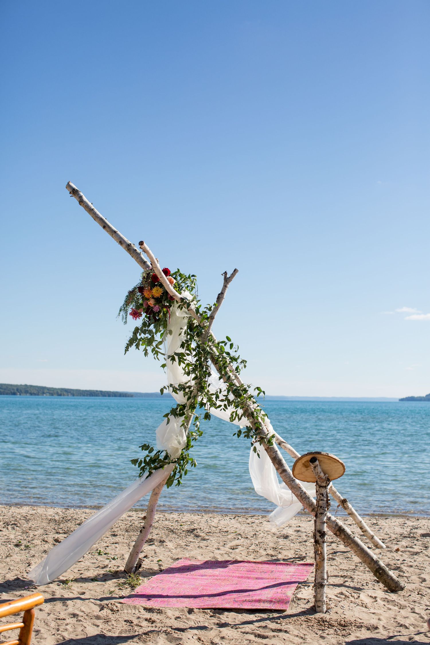 Rustic beach wedding arch made of driftwood and white fabric decorated with flowers along a turquoise shoreline.