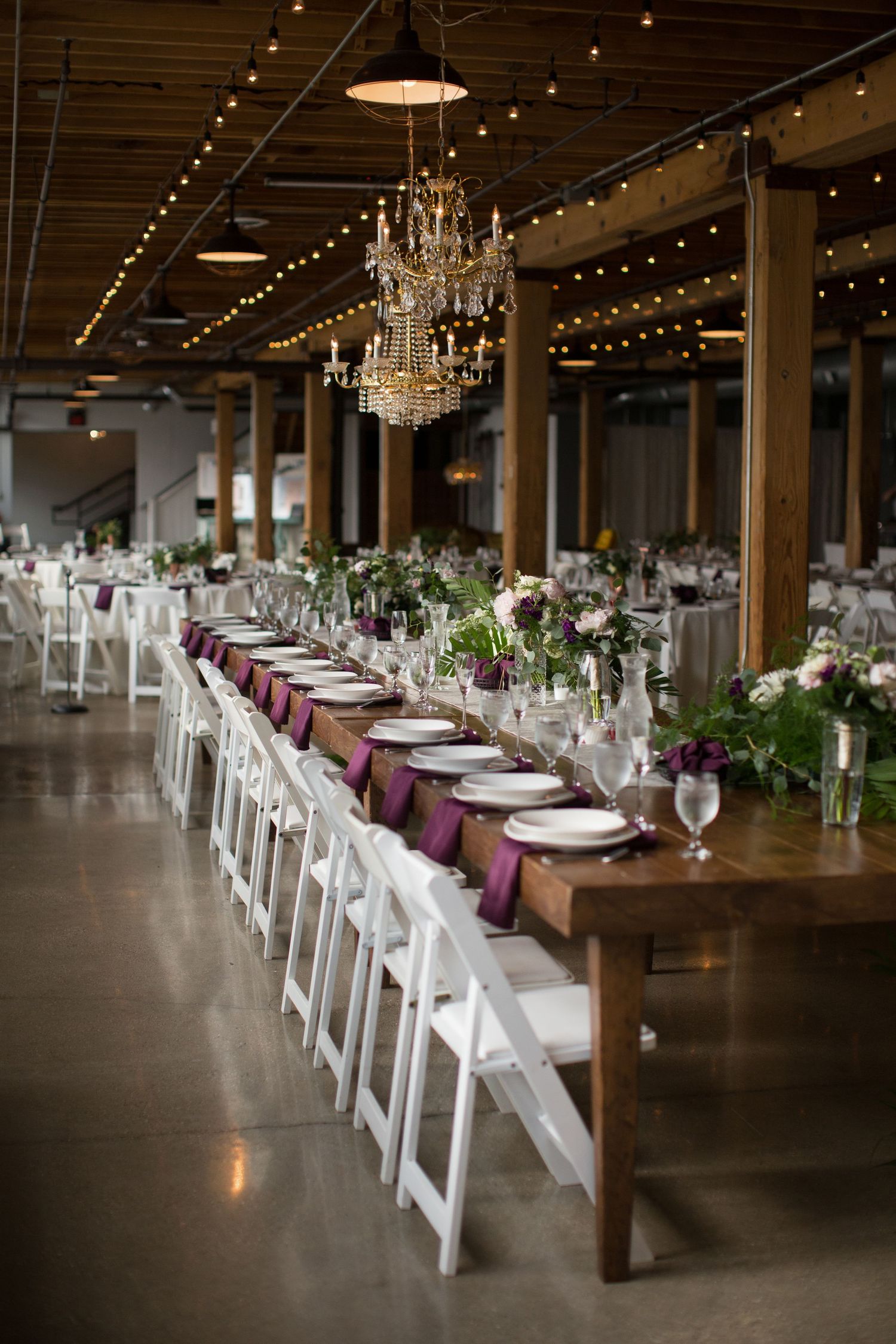 Long rustic wedding reception tables with white chairs and chandeliers in an industrial-style venue with string lights.