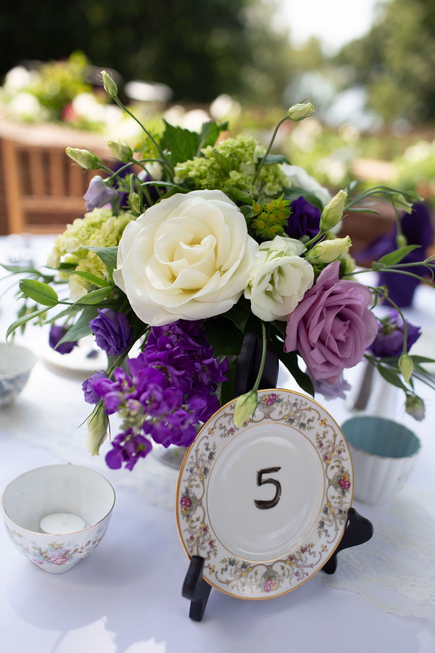 Elegant table settings with floral centerpieces featuring white roses and purple flowers alongside vintage china table numbers.