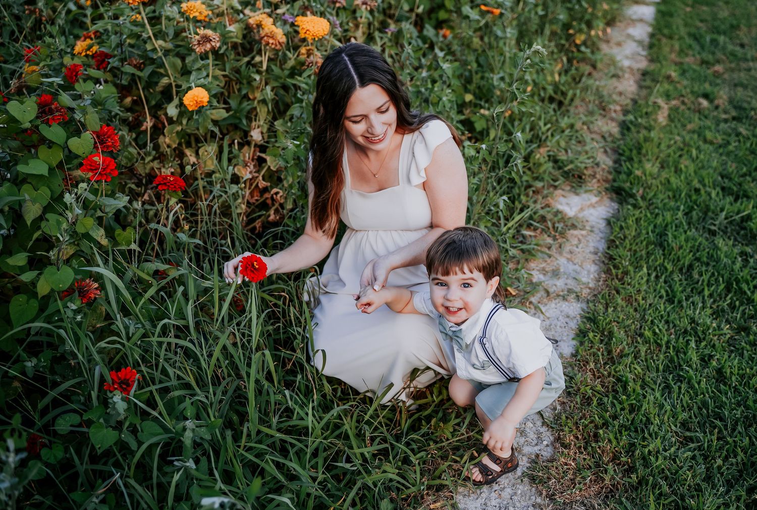 Two people kneeling on a garden path surrounded by blooming red and orange flowers on a sunny day.