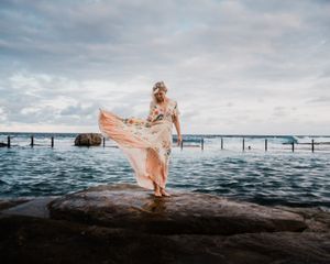 A flowing pink dress billows in the wind against a dramatic ocean backdrop at sunset.