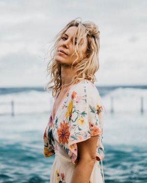 Woman in floral dress poses against ocean backdrop with crashing waves and cloudy sky.