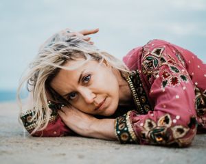 Person lying on sandy beach wearing pink floral dress and jewelry with ocean waves in background.