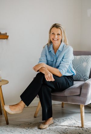 A person in a light blue shirt and dark pants sits casually on a modern gray armchair in a minimalist room.
