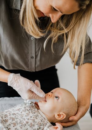 A caregiver wearing latex gloves administers medicine through a dropper to a baby lying on a floral blanket.