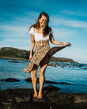 Woman in casual attire explores rocky coastline with blue ocean waters and island views in background.