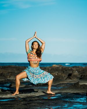 A person in a blue dress strikes graceful dance poses on dark volcanic rocks by the ocean at sunset.