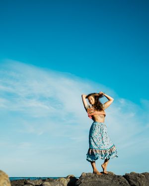A person in a blue floral dress stands on coastal rocks against a vibrant turquoise sky in a panoramic series.