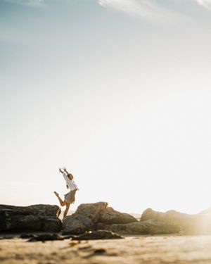 A silhouetted figure performs a graceful leap against a bright sunset sky over a rocky coastal landscape.