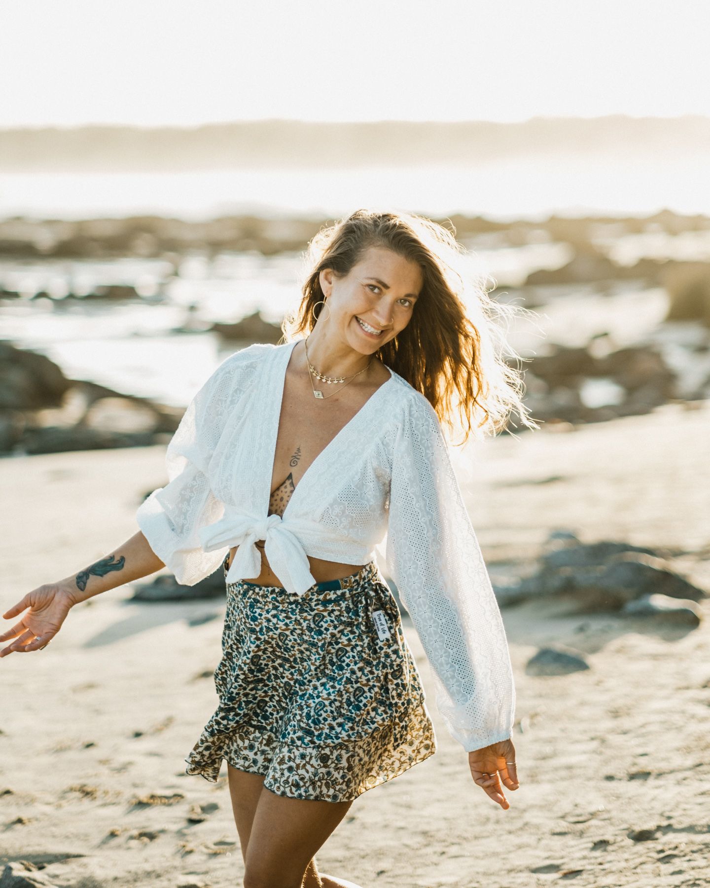 Woman in white tied top and printed skirt twirls on sandy beach at sunset.