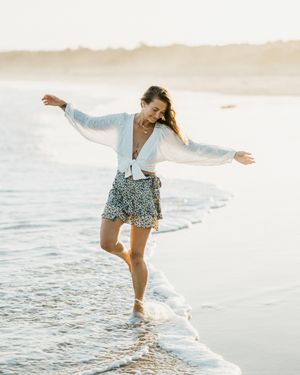 Woman in flowing white outfit dances joyfully along the shoreline at sunset.