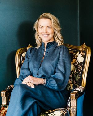 A person wearing a navy blue tunic sits in an ornate gold chair against a dark background.