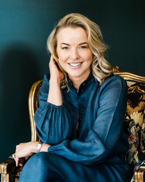 Professional portrait of someone in a navy blue blouse seated in an ornate vintage chair against a dark teal background.