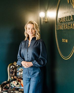 A person in a navy jumpsuit poses in front of a dark green wall with gold signage in a retail setting.