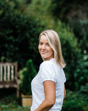 Person in a white t-shirt smiling while standing in a garden with a wooden bench in the background.