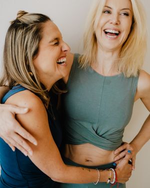 Two women laughing together and embracing while wearing casual workout attire in a bright indoor setting.