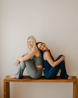 Two people in casual attire sit back-to-back on a wooden bench in a minimalist setting.
