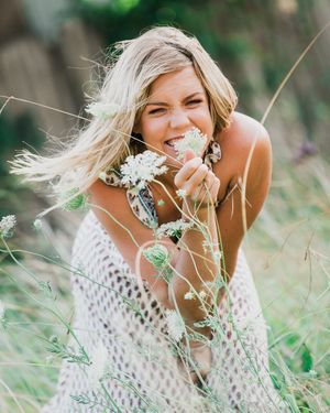 A person in a white crochet sweater smiles while smelling wildflowers in a sunny meadow with wind-blown hair.