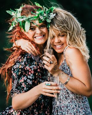 Two people wearing floral dresses and green leaf crowns embrace joyfully in a dark outdoor setting.