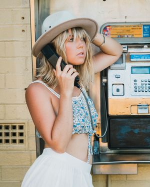 A person in a floral top and white skirt uses a vintage payphone while wearing a beige wide-brimmed hat.