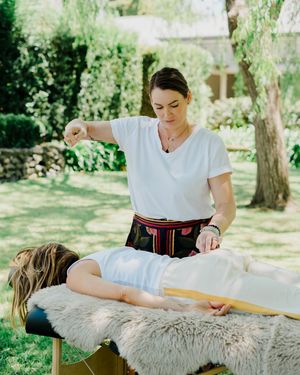 Massage therapist performs outdoor treatment on massage table covered with sheepskin in sunny garden setting.