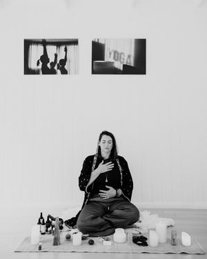 A person in dark clothing sits cross-legged on the floor with candles and small objects arranged in front of them.