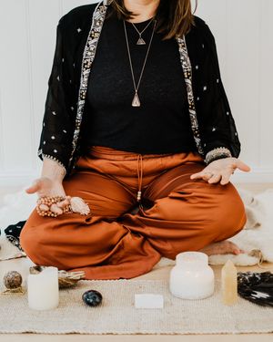 Person in black sweater and rust-colored pants sits cross-legged on floor with crystals and candles for meditation practice.