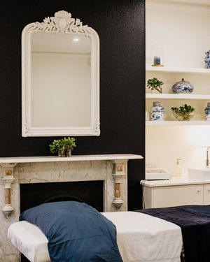 A spa treatment room with built-in white shelving, ornate mirror, and navy blue treatment bed.