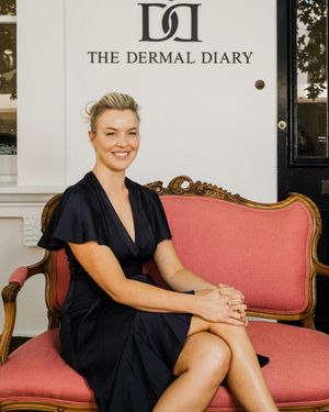 A person in a black dress sits elegantly on a pink vintage sofa in front of The Dermal Diary business sign.