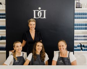 Four staff members pose together in front of a black wall with a company logo and blue shelving in the background.