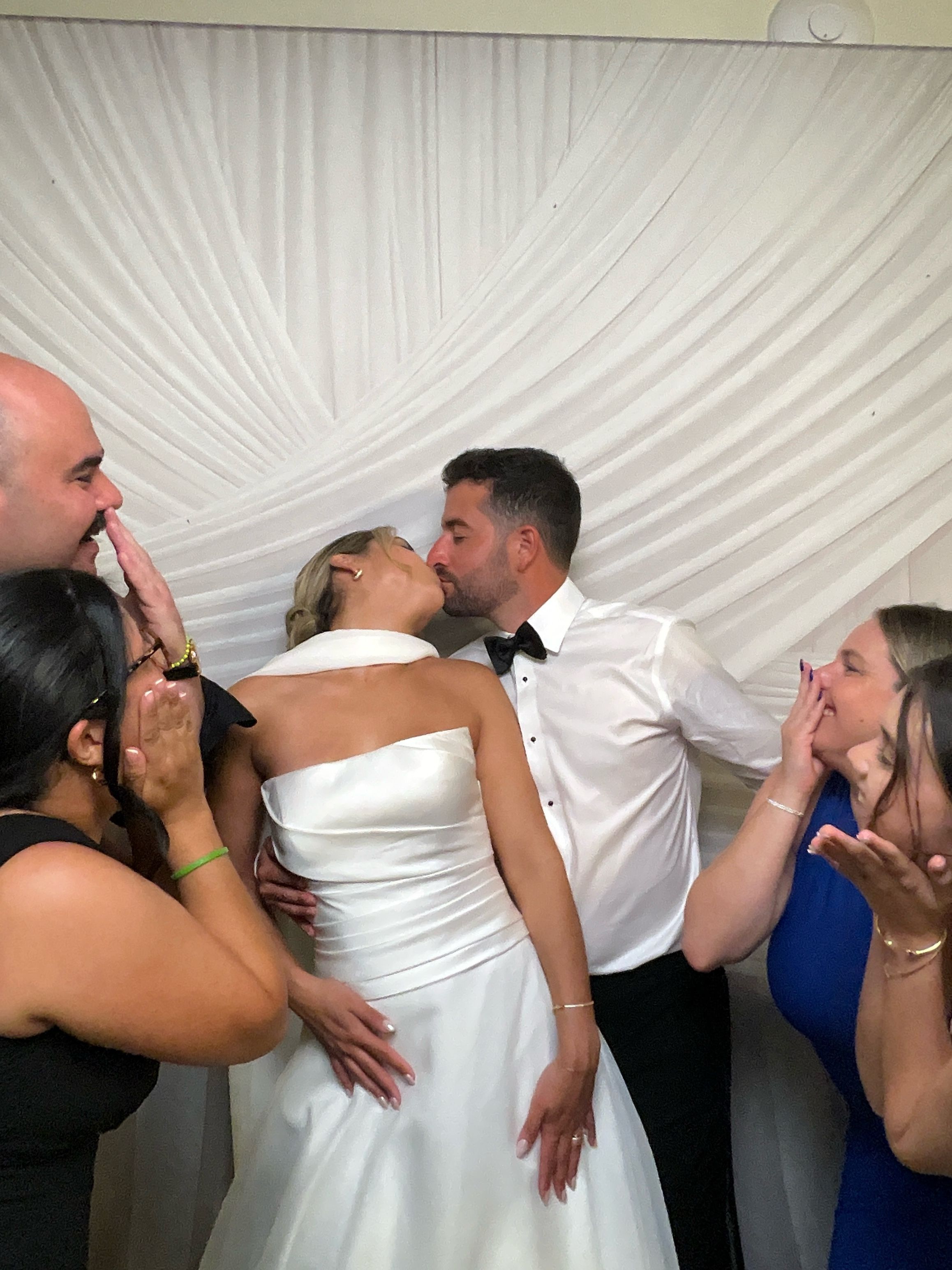 Bride and groom share a kiss at wedding reception while guests playfully pose beside them in front of white draped backdrop.