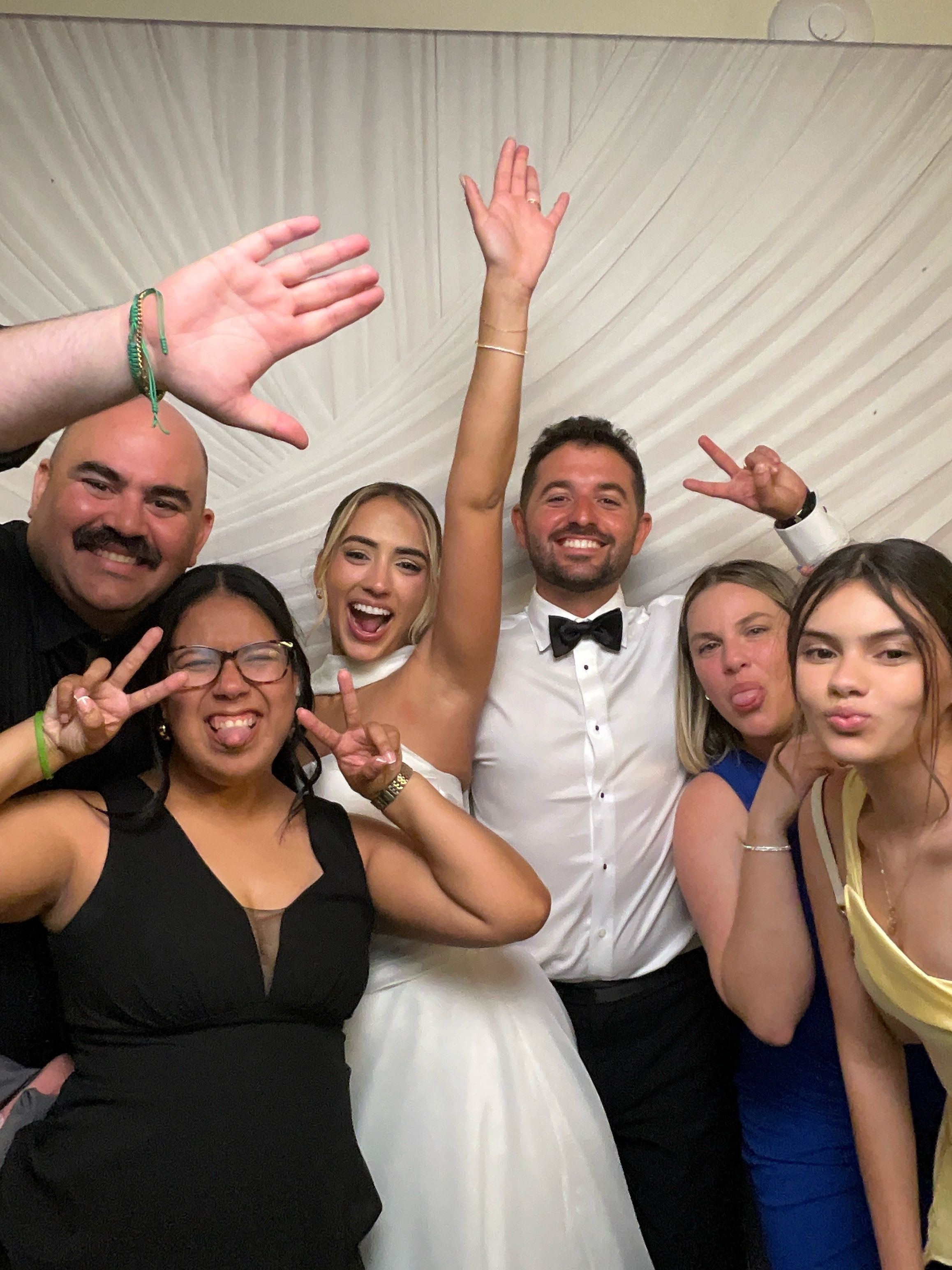 A joyful wedding group photo with bride in white dress, groom in tuxedo, and four friends making fun poses.